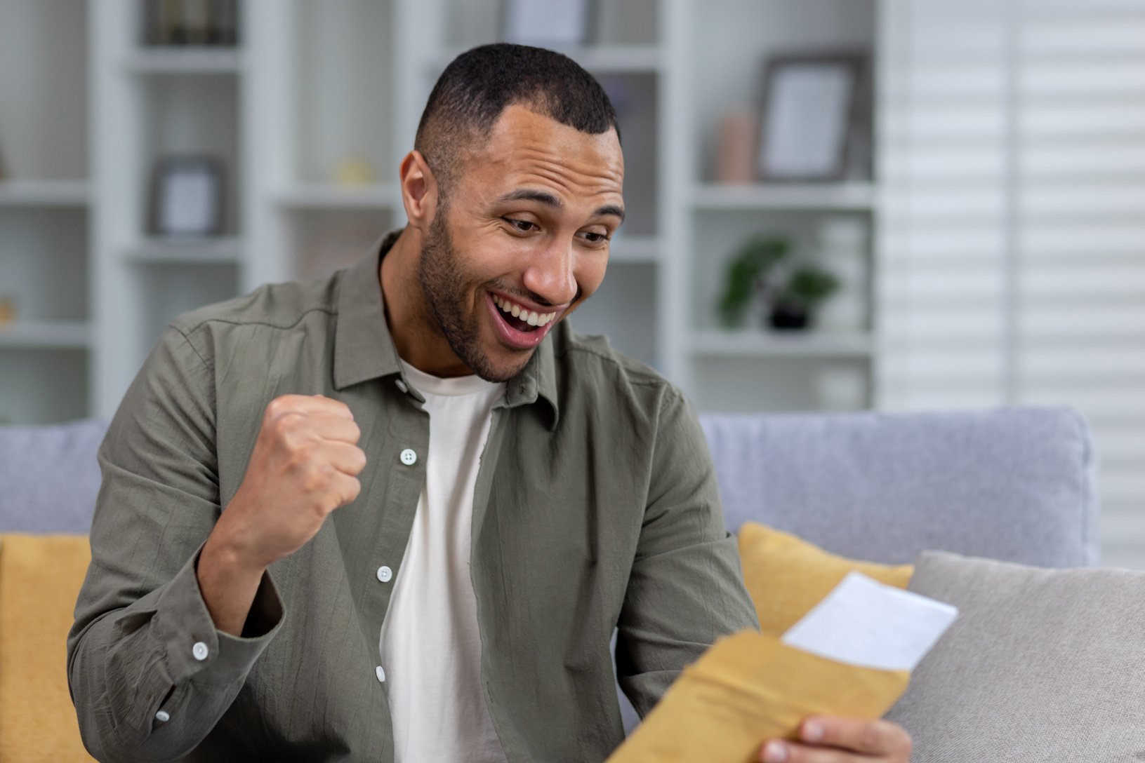 Happy hispanic young male student is happy to receive news. Sits at home on the couch and reads letters, documents, exam results, tests. Shows a victory gesture with his hand